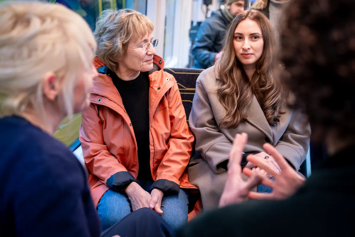 In einer Straßenbahn sitzen drei Frauen und eine vierte Person. Eine der Frauen und die vierte Person sieht man von hinten. Zwei Frauen schauen in Richtung der Kamera und sprechen mit den beiden anderen Personen. Gespraech_in_der_Bahn_mittel.jpg