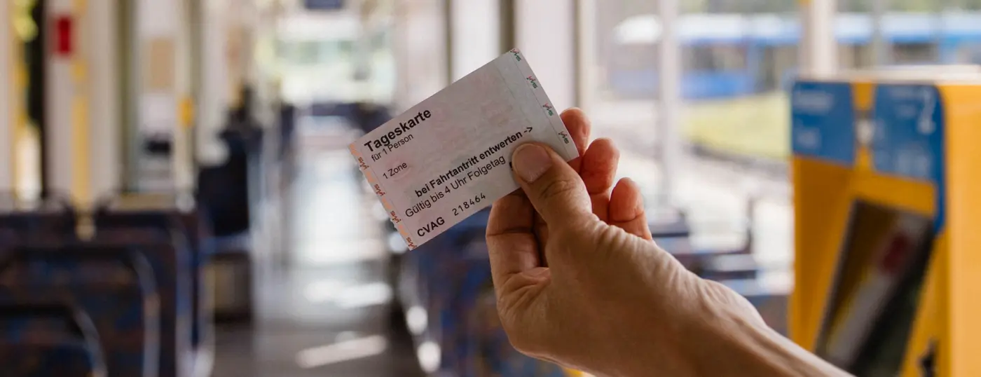 Eine Hand hält ein Ticket ins Bild. Im Hintergrund sieht man den Innenraum einer Straßenbahn.
