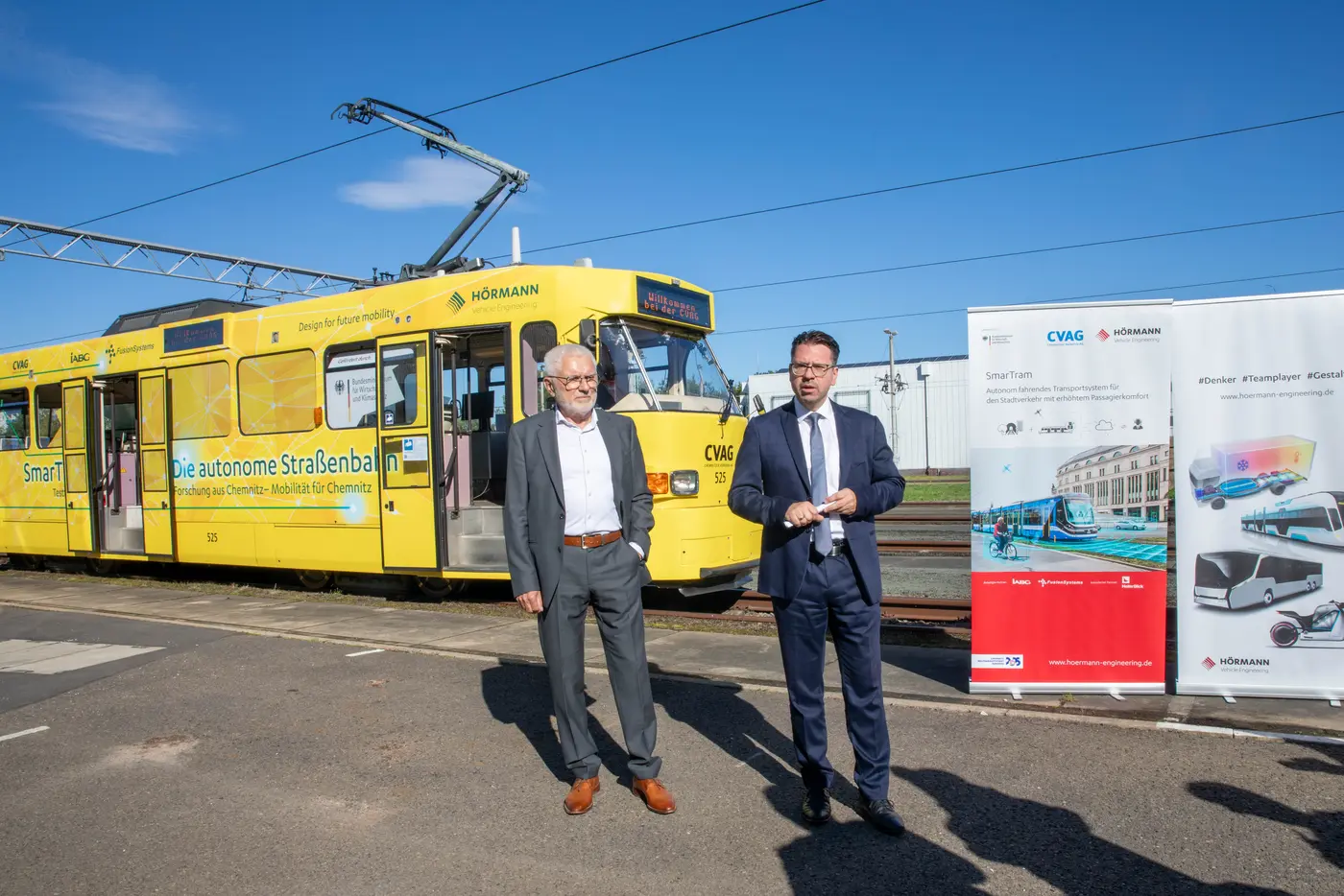 Dr. Volkmar Vogel, Senior Vice President vom Projektkonsortialführer HÖRMANN Vehicle Engineering (links im Bild) und David Joram, Geschäftsbereichsleiter Technik bei der CVAG (rechts im Bild) stehen vor der SmarTram. Neben ihnen steht ein Banner, welches die Logo der Projektpartner und eine Visualisierung einer Straßenbahn zeigt.