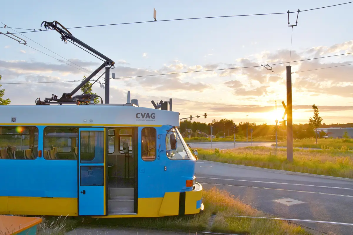 Eine Tatrabahn mit blau-gelber Lackierung steht an einer Haltestelle. Im Hintergrund sieht man den Sonnenuntergang.