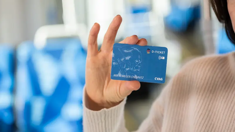 Eine Frau hält eine Chipkarte nah in die Kamera. Die Chipkarte ist blau. Auf der Chipkarte ist eine Skizze des Karl-Marx-Kopfes zu sehen. Außerdem sind das D-Ticket Logo, das CVAG Logo und der Spruch Wir bewegen Chemnitz abgebildet.